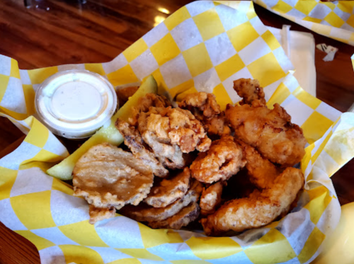 A basket of fried chicken pieces and pickles, served with a small cup of dipping sauce, on a checkered paper liner.