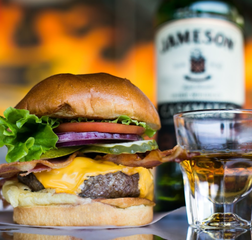 A juicy cheeseburger with lettuce, tomato, onion, and bacon, alongside a glass of whiskey and a bottle of Jameson.