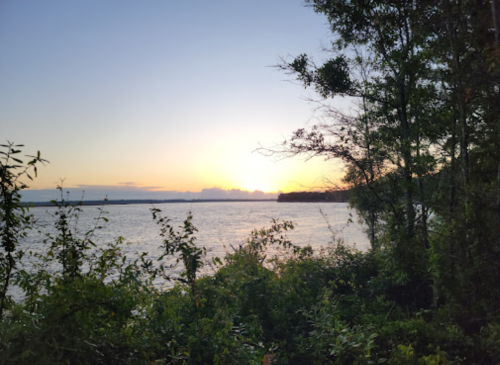 Sunset over a calm river, framed by lush greenery and trees along the shoreline.