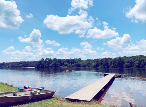 A serene lake scene with boats, a wooden dock, and fluffy clouds in a blue sky, surrounded by lush greenery.