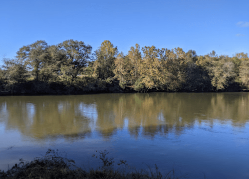 A calm river reflects trees and a clear blue sky, surrounded by autumn foliage.