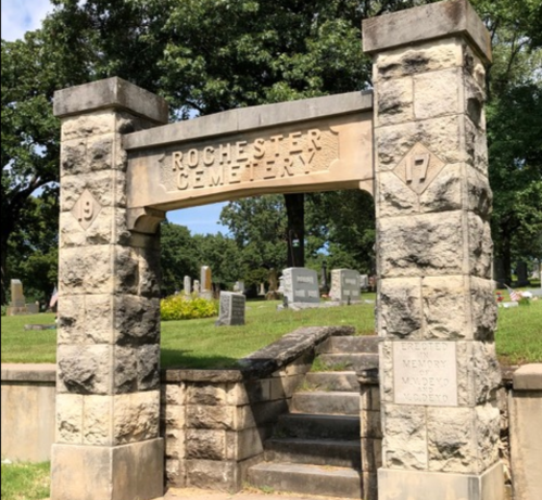 Stone entrance to Rochester Cemetery, featuring a sign and steps leading into a grassy area with gravestones.