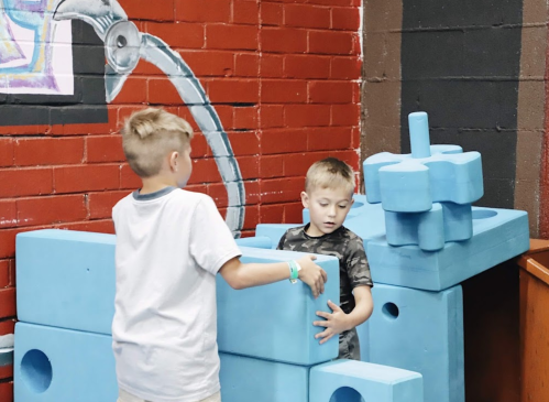 Two children play with large blue building blocks in a colorful indoor space.