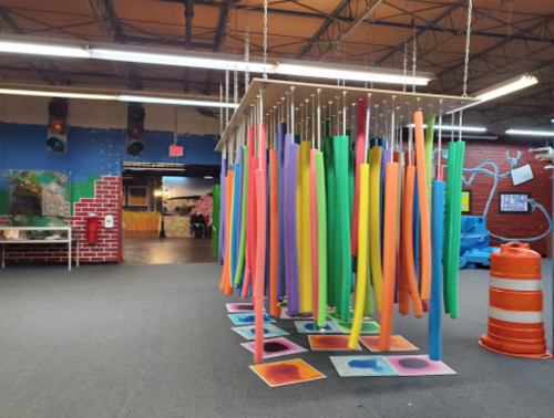 Colorful pool noodles hang from the ceiling in a spacious, vibrant indoor play area with art on the walls.