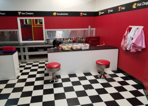 A colorful play kitchen with a checkered floor, featuring a counter, stools, and various food items displayed.