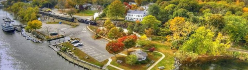 Aerial view of a scenic riverside park with autumn foliage, a train, and a docked boat.