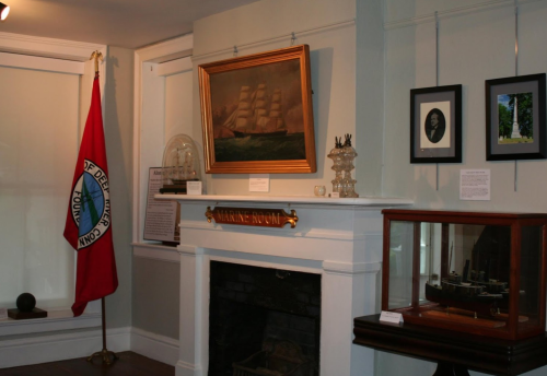 Interior of a museum room featuring a ship painting, a flag, framed photos, and a display case with a model ship.