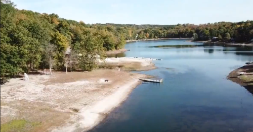 Aerial view of a serene lake surrounded by trees, with a sandy shore and a small dock extending into the water.