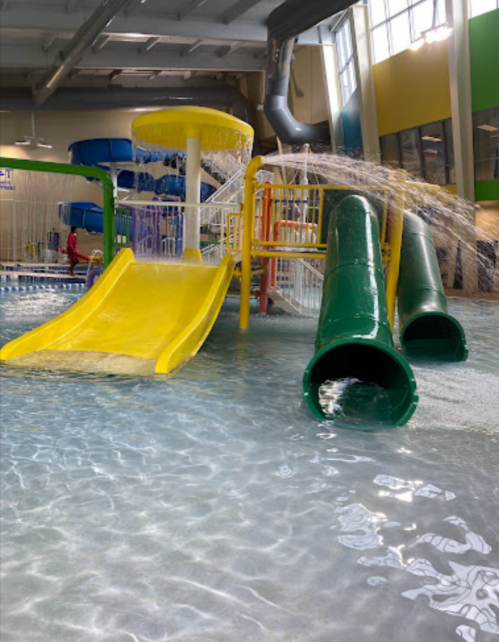 Colorful water slides and play structures in a bright indoor pool area with splashing water.