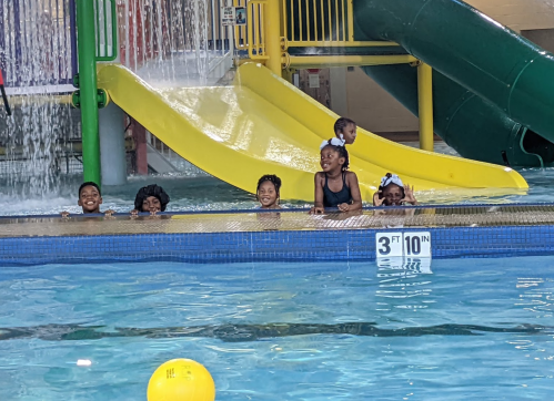 Children enjoying a water park, smiling in the pool near a yellow slide and a splash area.