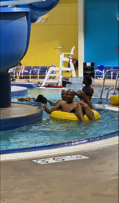 Two people relax on a yellow inner tube in a pool, with others swimming nearby and colorful walls in the background.