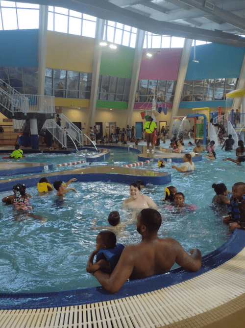 A lively indoor pool scene with children and adults enjoying the water, colorful walls, and play structures.