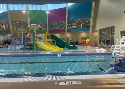 Indoor swimming pool with slides, lifeguard on duty, and people enjoying the water. Bright, colorful environment.