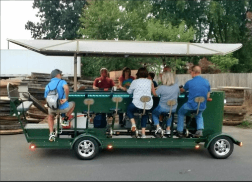 A group of people enjoying a pedal-powered bar on wheels, with a canopy, parked in a casual outdoor setting.