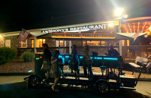 A group of people stands by a brightly lit trolley outside Anthony's Restaurant at night, with American flags displayed.