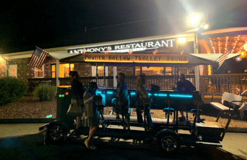 A group of people stands by a brightly lit trolley outside Anthony's Restaurant at night, with American flags displayed.