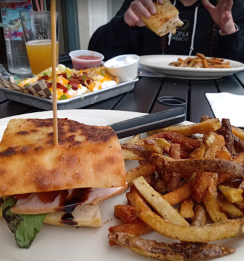 A close-up of a sandwich with fries on a table, with a person in the background holding food.