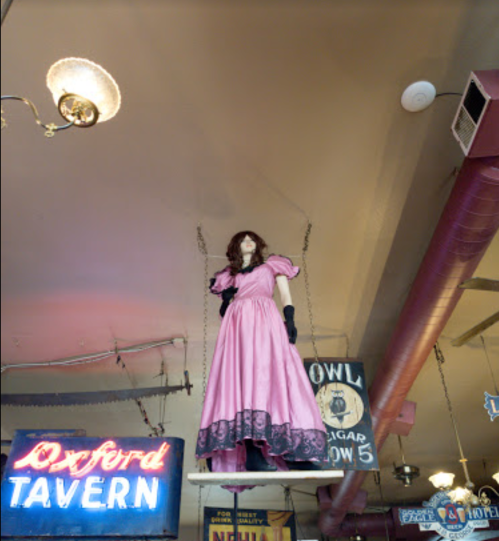 A vintage mannequin in a pink dress hangs from the ceiling in a tavern, surrounded by neon signs.
