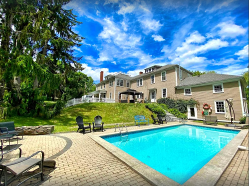 A sunny backyard with a swimming pool, patio furniture, and a large house surrounded by trees and blue skies.