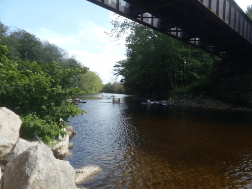 A serene river scene with kayakers paddling under a wooden bridge, surrounded by lush greenery and rocky banks.