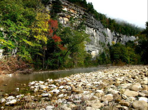 A serene river flows past rocky cliffs and colorful trees, shrouded in mist on a calm, overcast day.