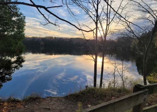 A serene lake at sunset, reflecting trees and clouds, with a wooden railing in the foreground.