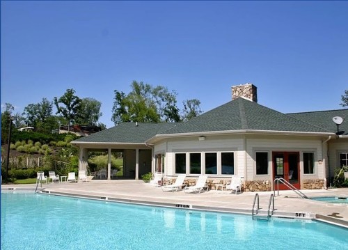 A sunny pool area with a building, lounge chairs, and trees in the background. Clear blue sky overhead.