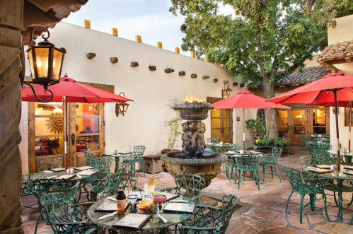 A charming outdoor dining area with green tables, red umbrellas, and a decorative fountain surrounded by lush greenery.