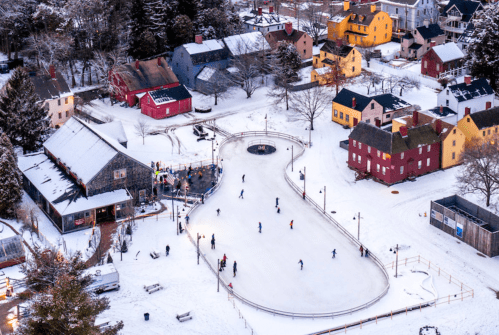 Aerial view of a snowy village with an ice skating rink and colorful houses, surrounded by trees.
