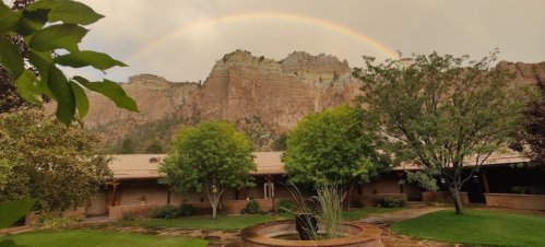 A serene courtyard with trees and a fountain, framed by a rainbow over majestic cliffs in the background.