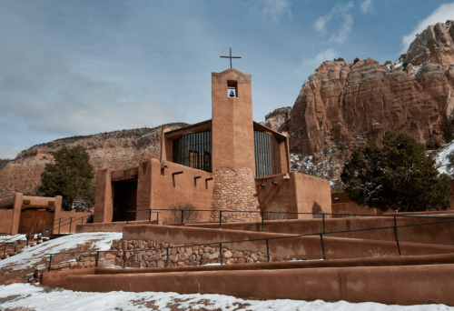 A modern adobe church with a cross, set against a backdrop of rocky cliffs and a snowy landscape.