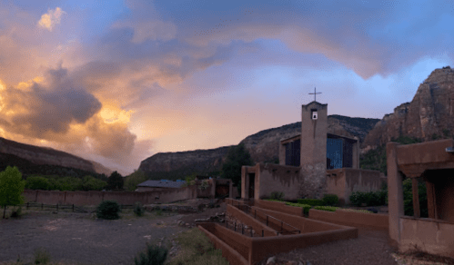 A scenic view of a church surrounded by mountains and dramatic clouds at sunset.