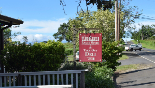 Sign for Ulupalakua Ranch Store & Take Out Deli, with a scenic view and a road in the background.
