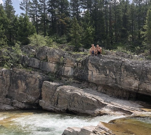 Two people sit on a rocky ledge by a flowing river, surrounded by lush green trees and a natural landscape.