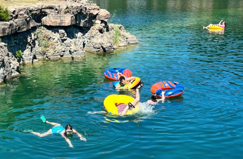 A sunny day at a lake with people swimming and floating on colorful inner tubes near rocky shores.