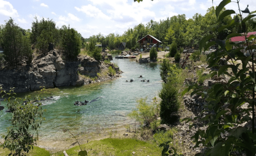 A serene river flows through a lush green landscape, with rocky banks and a few people enjoying the water.