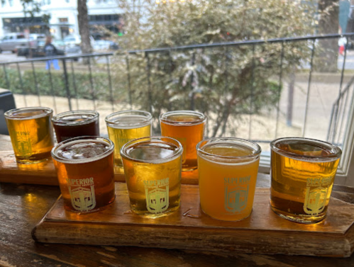 A wooden flight board with eight small glasses of various beers, set against a blurred outdoor background.