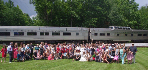 A large group of people poses together in front of a vintage train in a green outdoor setting.
