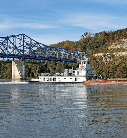 A riverboat passes under a blue bridge, surrounded by trees and a calm waterway on a clear day.