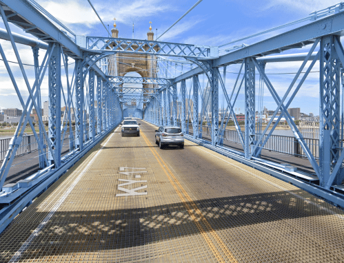 A blue steel bridge with cars driving on it, spanning a river under a clear sky.