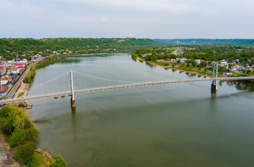 Aerial view of a suspension bridge over a river, surrounded by green hills and a small town along the shore.
