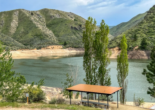 A serene lake surrounded by green mountains, with a shaded picnic area in the foreground.