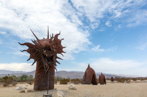 A large metal dragon sculpture stands in a desert landscape, with smaller structures in the background under a cloudy sky.