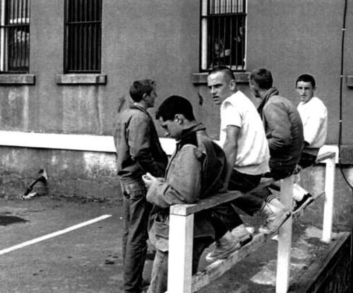 A group of young men sitting on a railing outside a building with barred windows, looking in different directions.