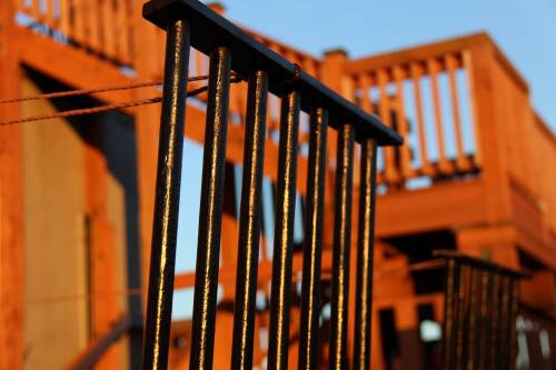 Close-up of black metal railings with a wooden structure and blue sky in the background, illuminated by warm sunlight.