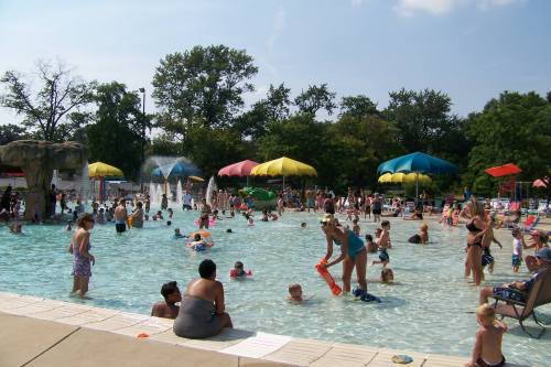 A busy outdoor pool scene with families and children enjoying the water under colorful umbrellas on a sunny day.