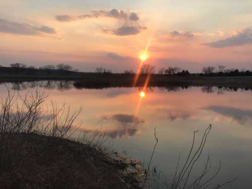 A serene sunset over a calm lake, with reflections of the sun and clouds in the water, surrounded by bare trees.