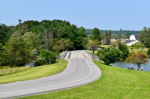 A winding road leads to a bridge over a calm river, surrounded by trees and grassy fields under a clear blue sky.