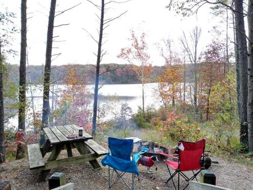 A serene lakeside campsite with a picnic table, two chairs, and autumn foliage reflecting in the water.