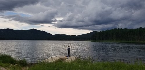 A person stands on a dock by a lake, surrounded by mountains and dark, cloudy skies.